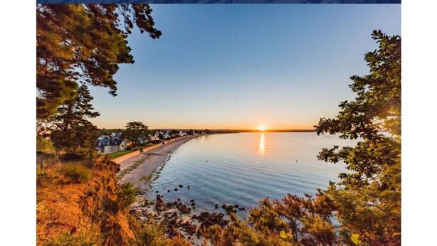 A coastal landscape with a sunset, calm water, and a row of houses along the shore.