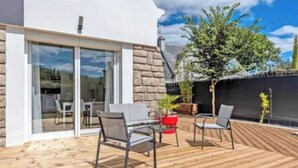 A modern outdoor patio with a wooden deck, a glass door leading to an interior space, and a small tree.