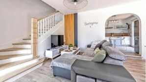 A modern living room with a staircase, a sofa, a TV, and a kitchen area visible through an archway.