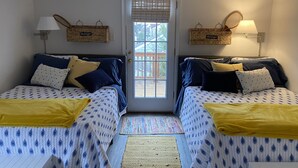 A room with two beds, each with a headboard, a small table, and a door. There are baskets on the wall and a window with blinds. - Ventnor newly renovated beach condo (Ventnor City)