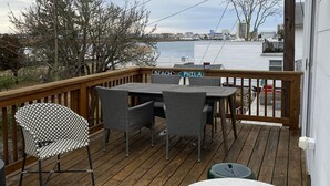 A wooden deck with a table and chairs, a round white table, and a view of a body of water and buildings in the background. - Ventnor newly renovated beach condo (Ventnor City)