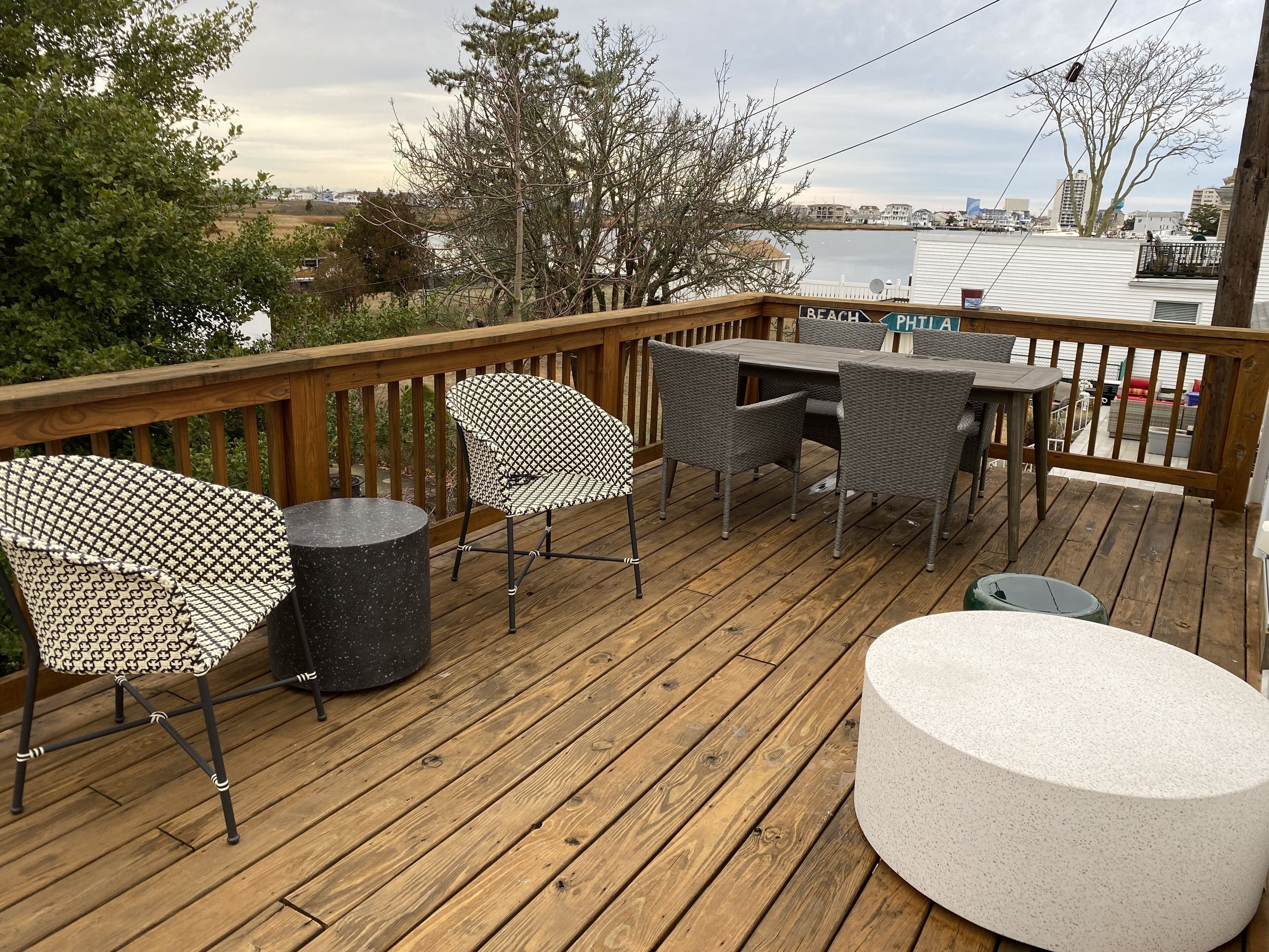 A wooden deck with outdoor furniture, including chairs and a table, overlooking a body of water and buildings in the distance.