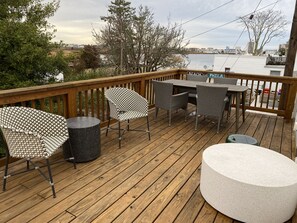 Une terrasse en bois, aménagée avec du mobilier d’extérieur, notamment des chaises et une table, d’où l’on peut admirer un plan d’eau et des bâtiments situés au loin.