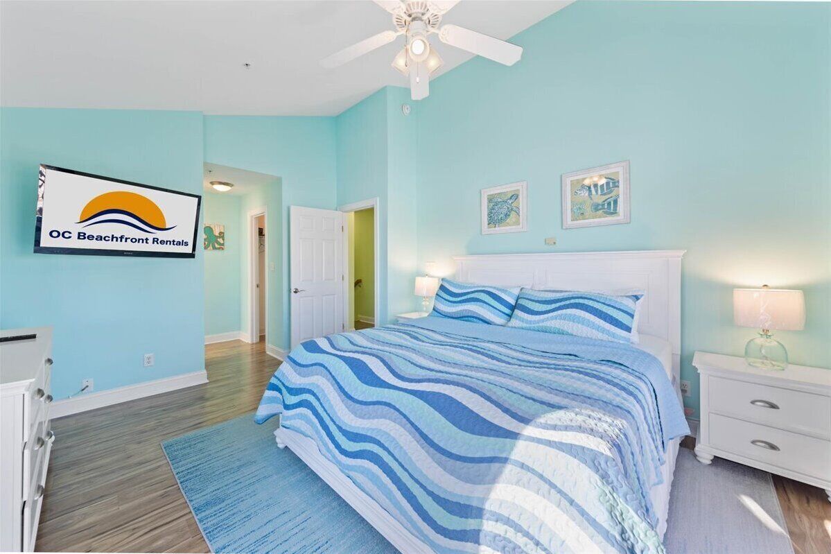 Une chambre avec un lit, une table de chevet, un ventilateur de plafond et un panneau indiquant « OC Beachfront Rentals ».