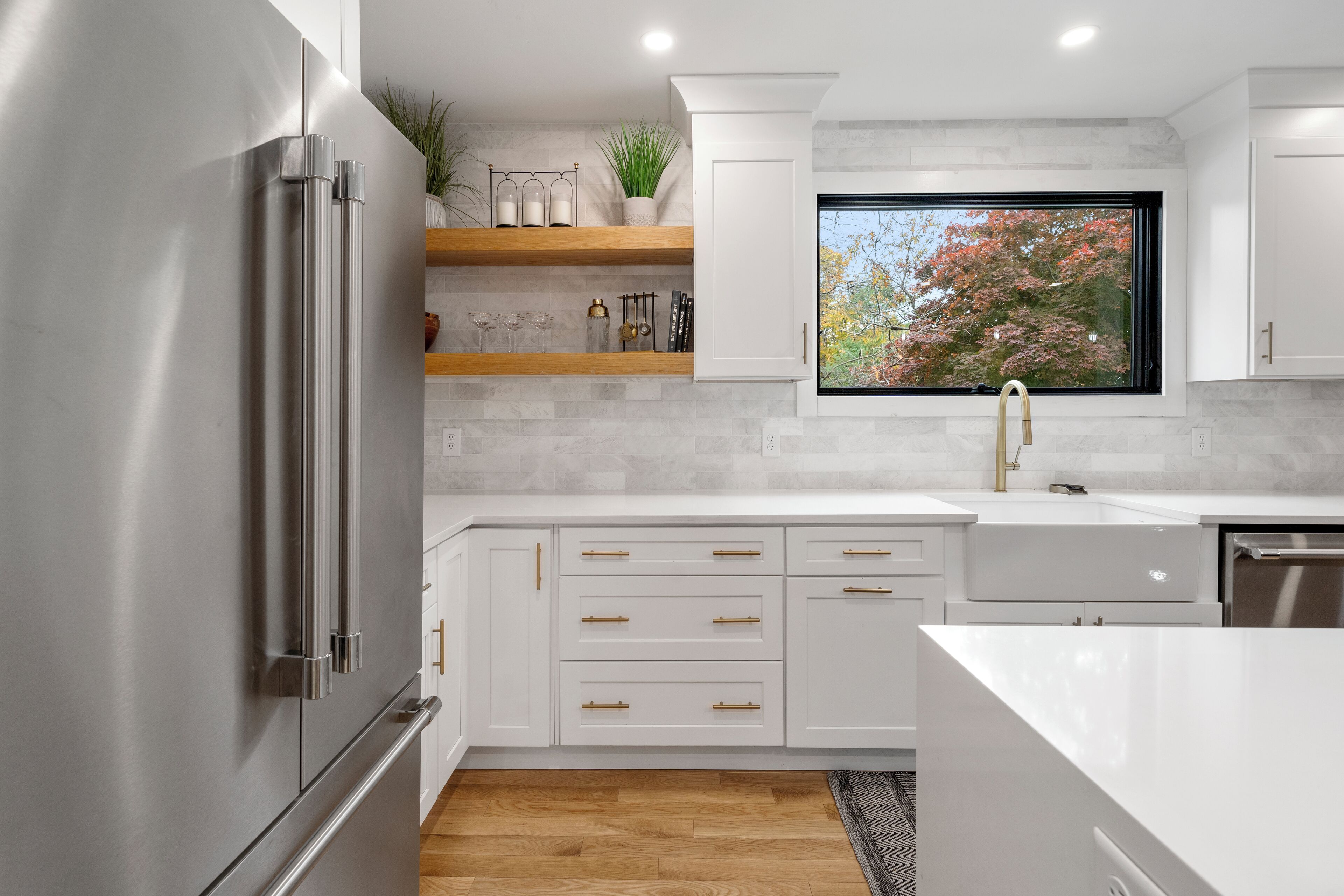 Una cocina moderna con armarios blancos, un refrigerador de acero inoxidable, estantes de madera y un ventanal con vista a árboles.