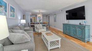 A living room with a grey sofa, a coffee table, a teal cabinet, and a TV on the wall. - Seasider 304 - Three Bedrooms, Three Bath Condo in Oceanfront Complex (Garden City Beach)