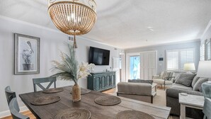 A modern living room with a wooden dining table, a sofa, and a television. - Seasider 304 - Three Bedrooms, Three Bath Condo in Oceanfront Complex (Garden City Beach)
