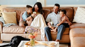 A family on a brown leather couch with a dog, a vase of flowers, and a tray with food items.