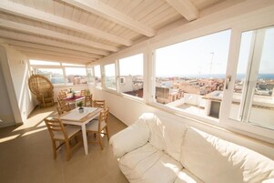 Una stanza luminosa con soffitto in legno, un divano bianco, un tavolo da pranzo con sedie e ampie finestre che offrono vista su edifici e mare.