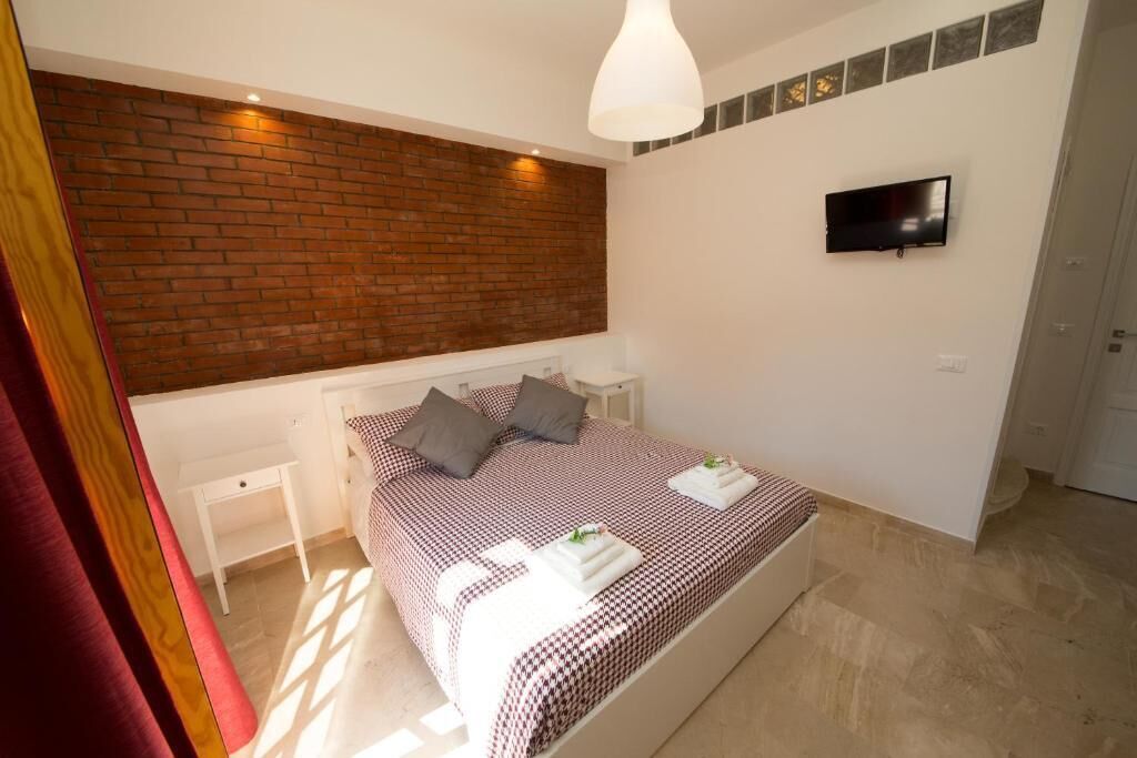 A bedroom with a brick accent wall, a bed with a checkered bedspread, a flat-screen TV mounted on the wall, and a small bedside table.
