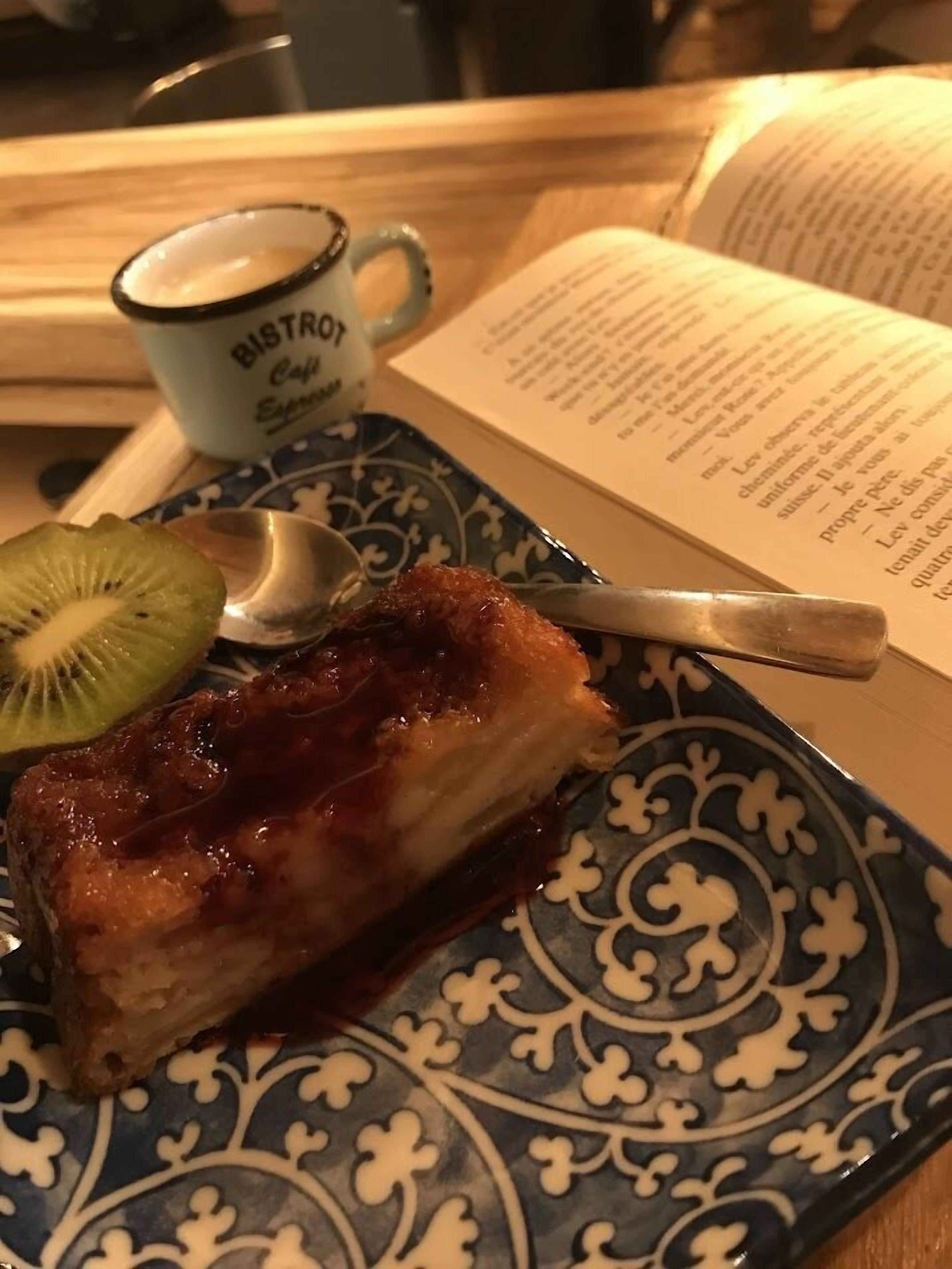A dessert plate with a slice of cake, a kiwi slice, and a spoon, accompanied by a coffee cup and an open book.