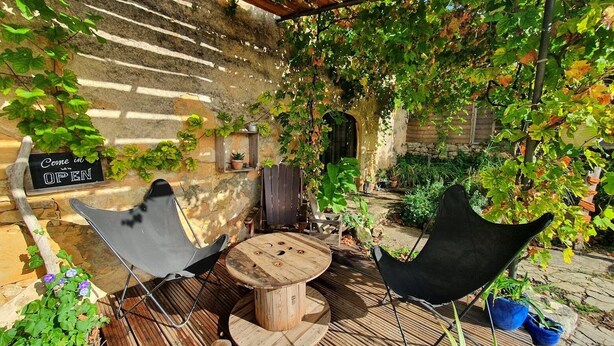 An outdoor seating area with a wooden table and two chairs, a sign that says "Come in OPEN", and a stone wall with climbing plants.