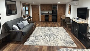 A modern living room with a dark-colored sofa, a large patterned rug, wooden flooring, and a kitchen area with wooden cabinets and appliances.
