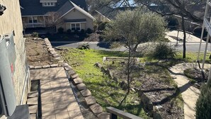 A residential area with a paved pathway leading to a house with a porch, surrounded by trees and a view of a lake.