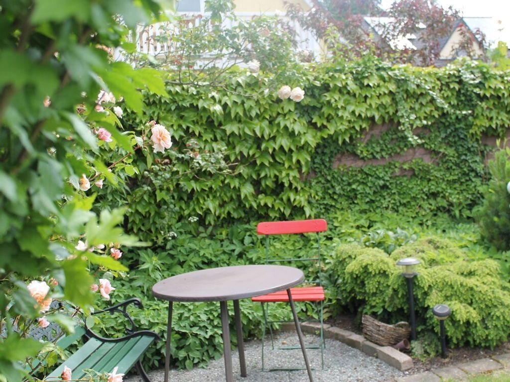 Un jardín con una mesa redonda y dos bancos rojos, rodeado de follaje verde exuberante y plantas en flor.