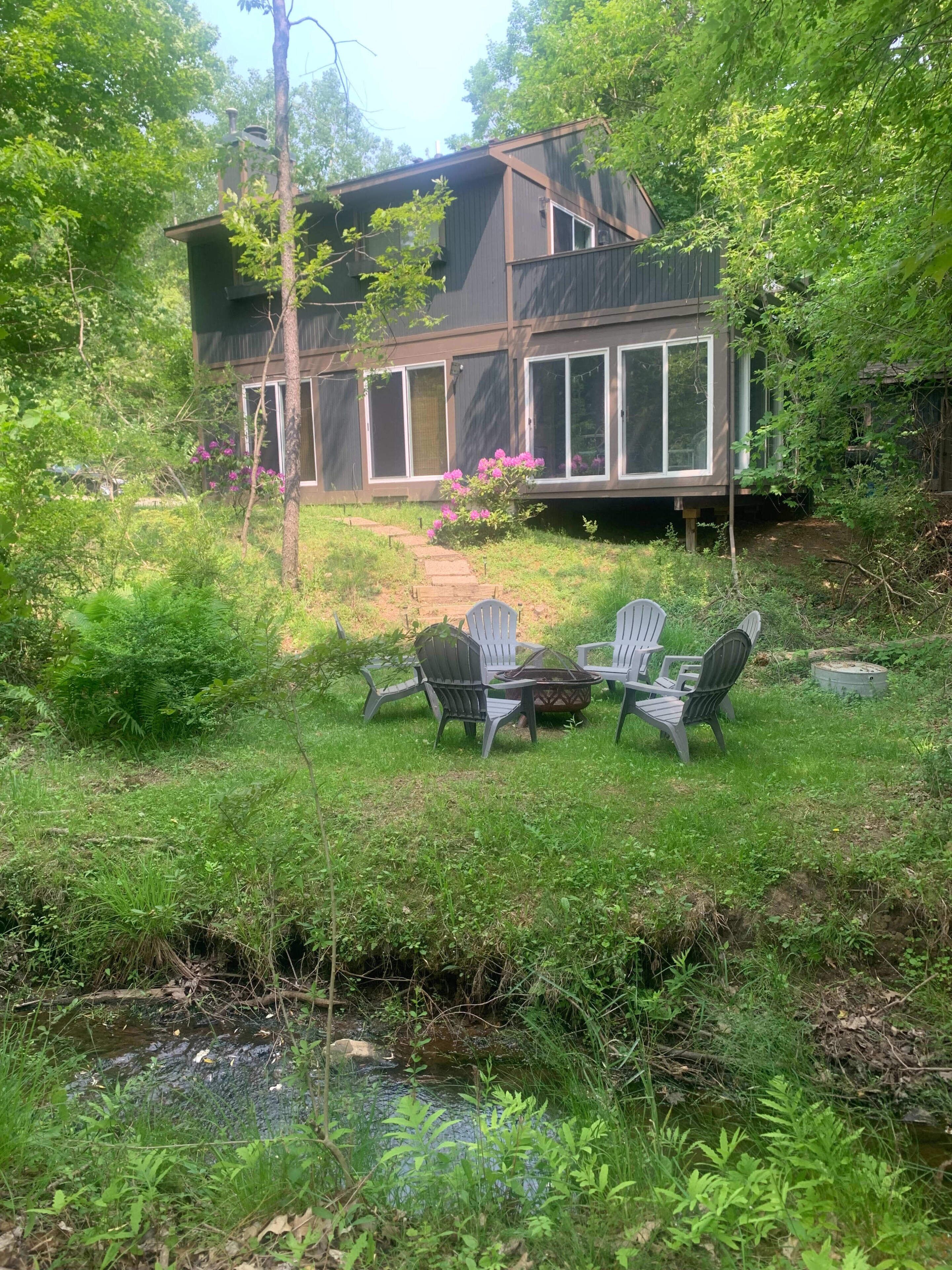 Et hus med terrasse, omgivet af frodig vegetation og en lille å.