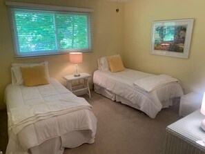 A bedroom with two single beds, a nightstand with a lamp, a window with blinds, and a framed picture on the wall. - Whittier Cottage (Douglas)