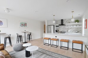 Een moderne keuken met witte kasten, een marmeren spatwand en een zwarte afzuigkap. Drie barkrukken met leren zittingen en metalen frames, en een wit kookeiland met een spoelbak.