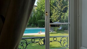 A window with a view of a swimming pool, green lawn, and a patio area. - Logis des Tourelles (Cognac)