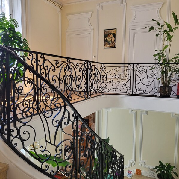 A staircase with ornate black metal railings, a framed picture on the wall, and potted plants. - Logis des Tourelles (Cognac)