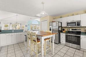 A modern kitchen with white cabinets, a wooden dining table, and stainless steel appliances. - Beach Bound: Private Pool | Hot Tub | Rec Room (Southern Shores)