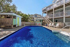 A swimming pool with a wooden deck, a small gazebo, and a multi-story building with balconies. - Beach Bound: Private Pool | Hot Tub | Rec Room (Southern Shores)