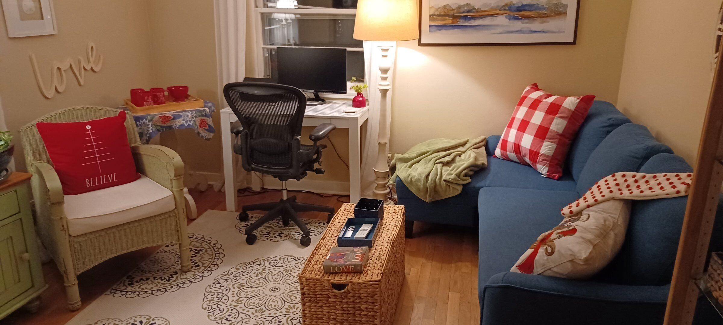 A cozy living room with a blue sofa, a wicker chair, a desk with a computer, and a bookshelf.