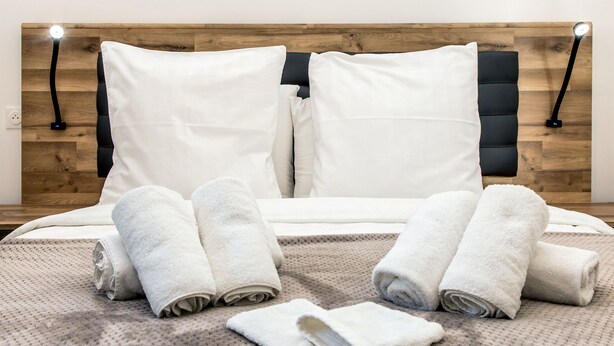Een net opgemaakt bed met witte kussens en opgerolde handdoeken, een houten hoofdbord en twee bedlampjes.