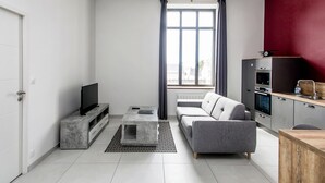 Een moderne woonkamer met een grijze bank, een flatscreen televisie op een marmeren console, een houten eettafel en een keuken met een rode accentmuur en roestvrijstalen apparatuur.