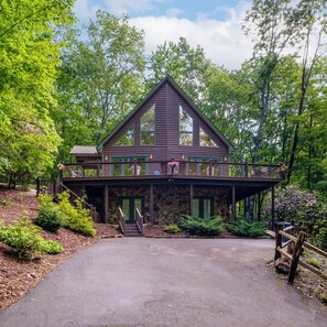 A two-story cabin with a stone foundation, a balcony with outdoor seating, and a driveway leading to the entrance.