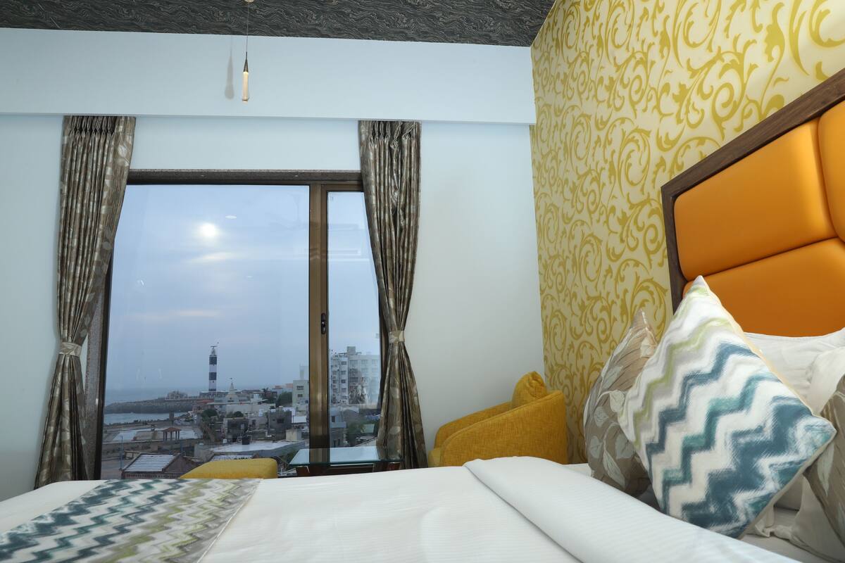 Quarto de hotel com uma janela grande, cama com roupa de cama estampada e vista para a paisagem urbana.