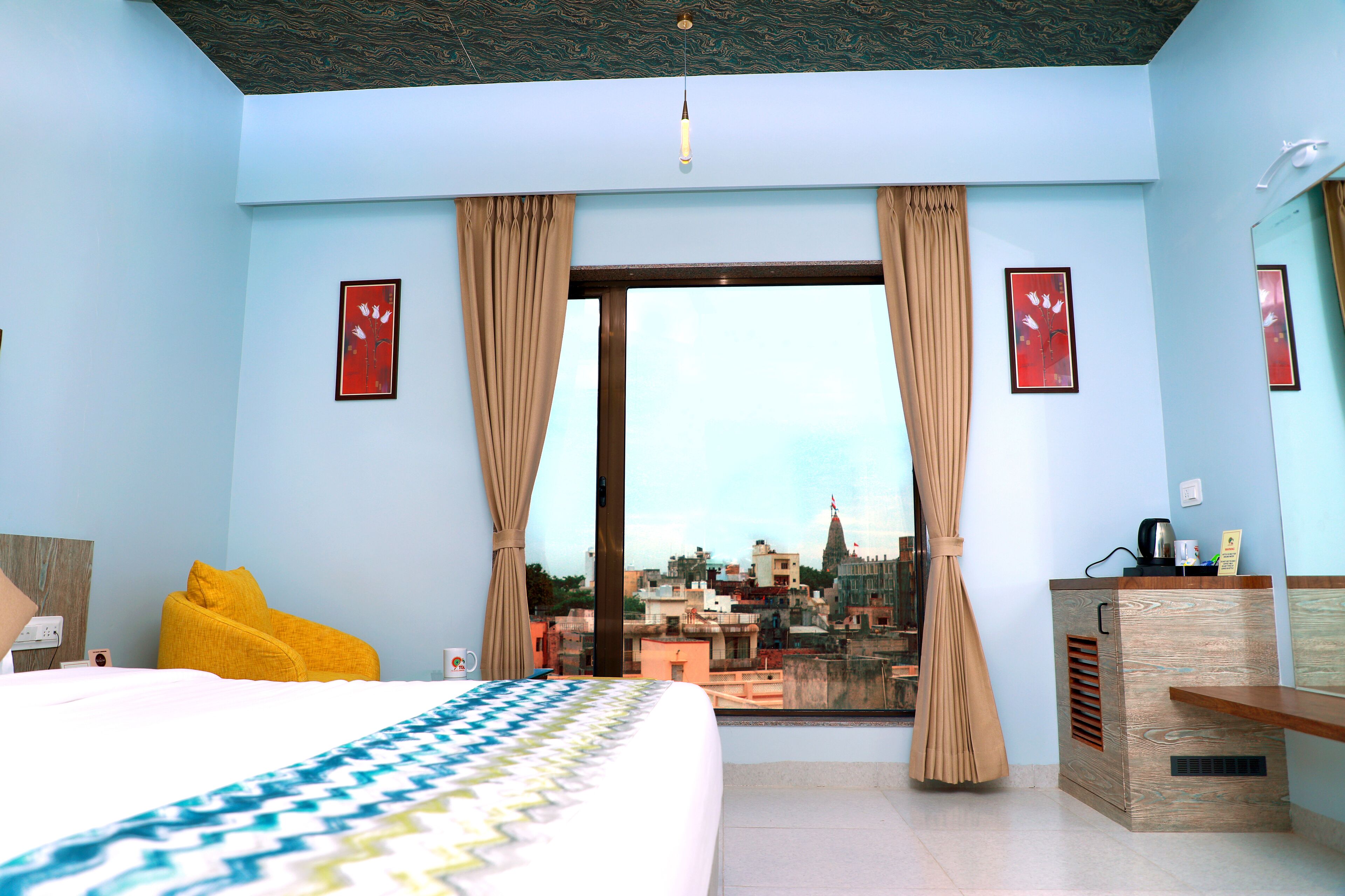 Habitación de hotel con cama, ventana con cortinas, vistas a la ciudad, escritorio con espejo y una pequeña mesa con hervidor.
