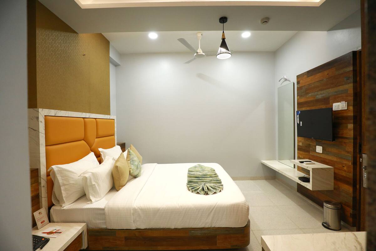 Quarto de hotel moderno com cama, cabeceira de madeira, TV fixada na parede e uma pequena mesa de cabeceira.