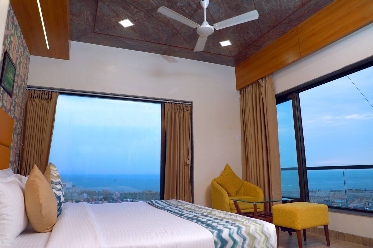 Quarto de hotel com uma cama grande, uma poltrona amarela e vista para o mar.