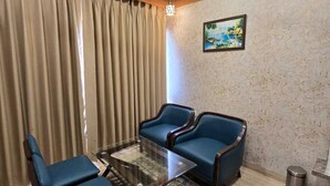 A room with a ceiling fan, a glass coffee table, two blue armchairs, a patterned rug, and a painting on the wall. - Hotel The Grand Ladhukara (Dwarka)
