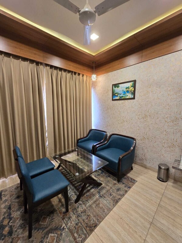 A room with a ceiling fan, a glass coffee table, two blue armchairs, a patterned rug, and a painting on the wall. - Hotel The Grand Ladhukara (Dwarka)