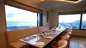 A dining area with a long table, cushioned seating, and large windows offering a city view. - Hotel The Grand Ladhukara (Dwarka)