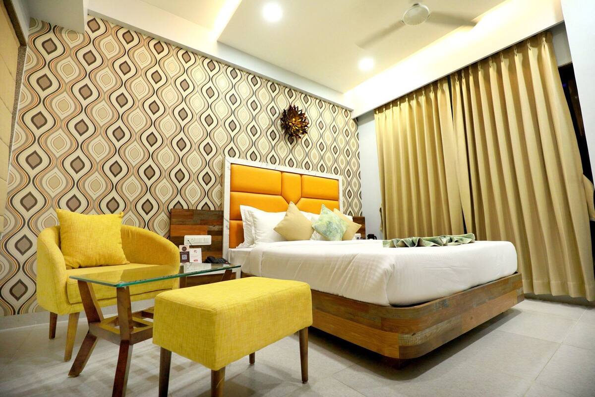 Quarto de hotel moderno com uma cama, uma mesa de centro de vidro, uma poltrona amarela e papel de parede estampado.