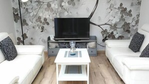 A modern living room with a white sofa, a coffee table, a TV, and a floral wall design. - Gîte en Sologne - La Deniserie + Garage (Romorantin-Lanthenay)