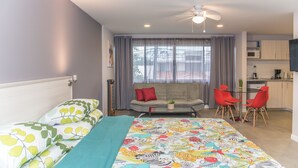 Un dormitorio con cama, sofá, zona de comedor con sillas, ventilador de techo y cocina.