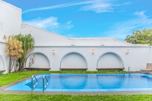 A swimming pool with a blue edge, surrounded by a white building with arched windows and a tiled roof.