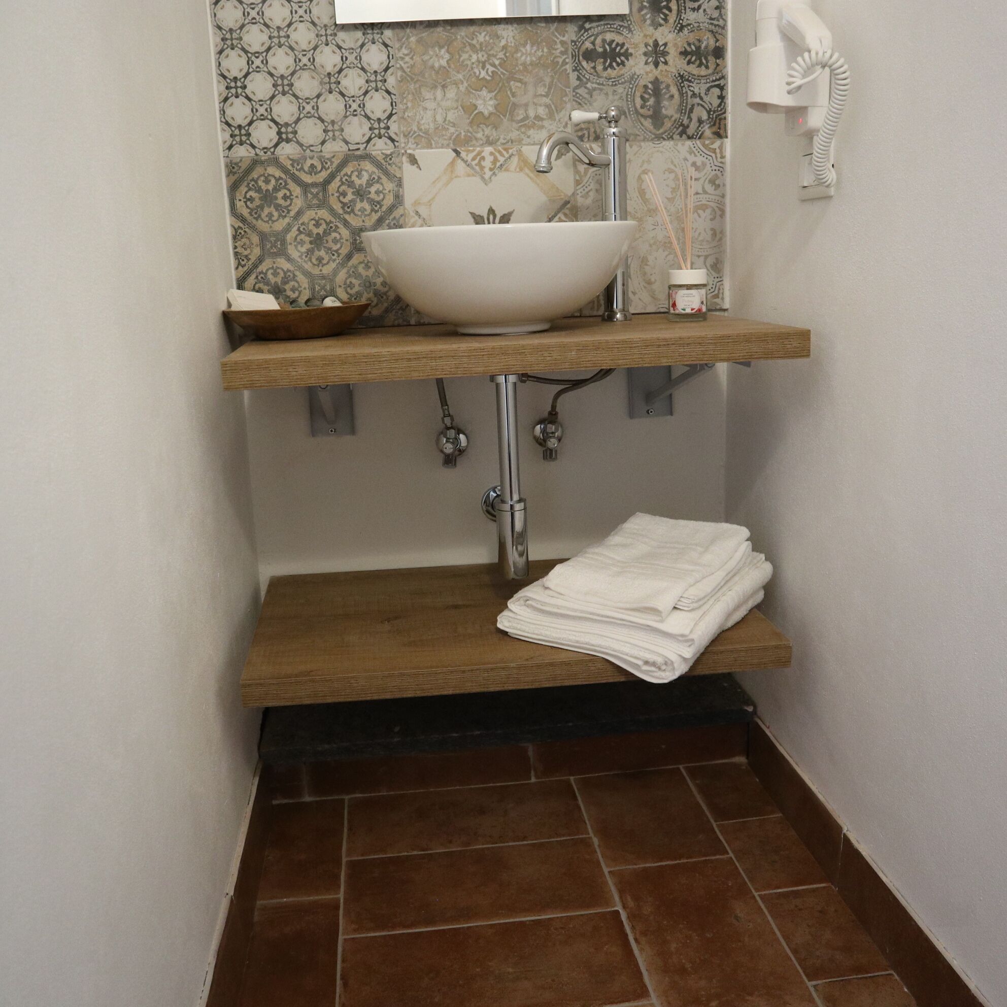A bathroom with a white vessel sink, a wooden shelf, and a towel stack.