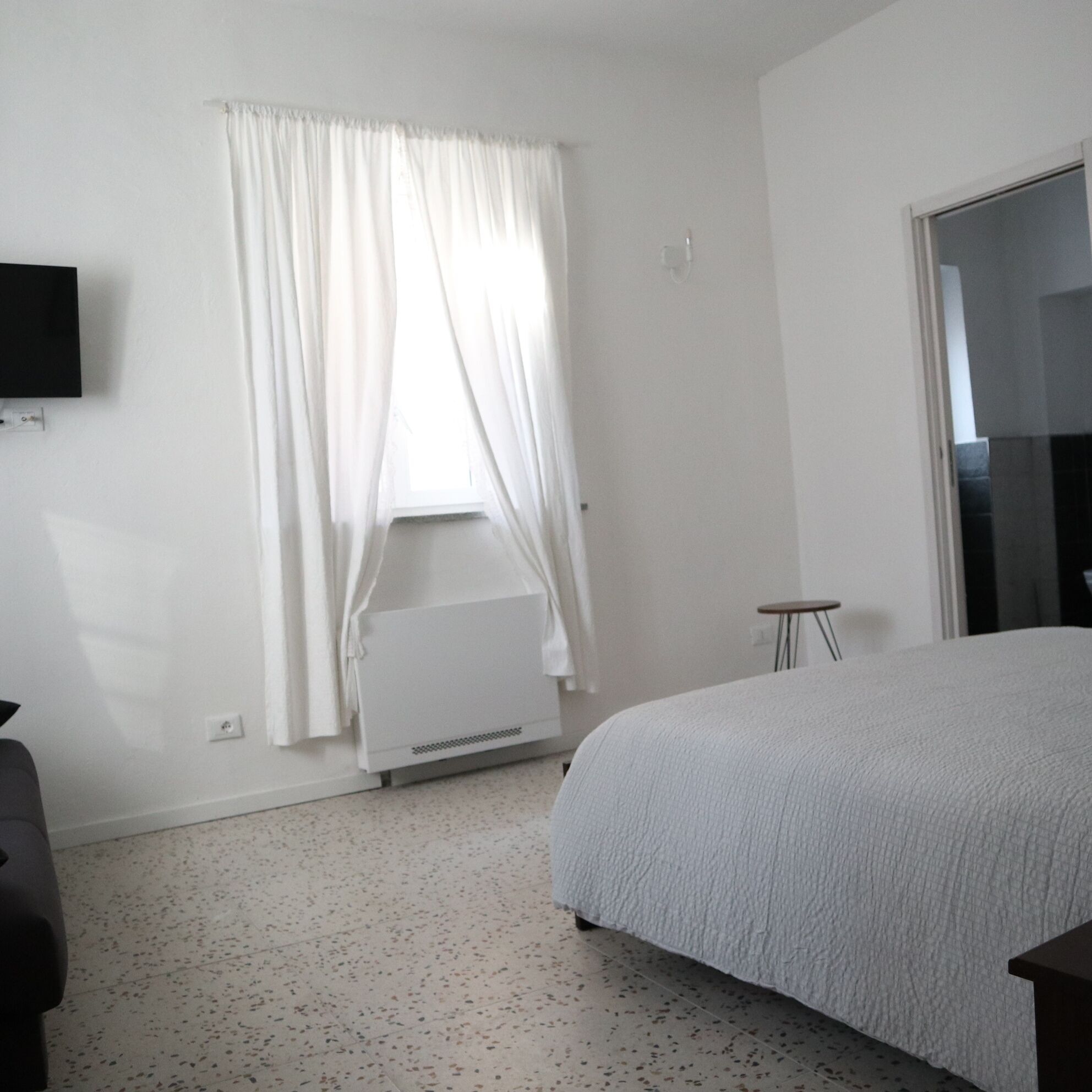 Une chambre à coucher avec un lit, une fenêtre avec des rideaux, un radiateur, une petite table et un téléviseur fixé au mur.