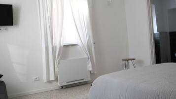 Une chambre à coucher avec un lit, une fenêtre avec des rideaux, un radiateur, une petite table et un téléviseur fixé au mur.