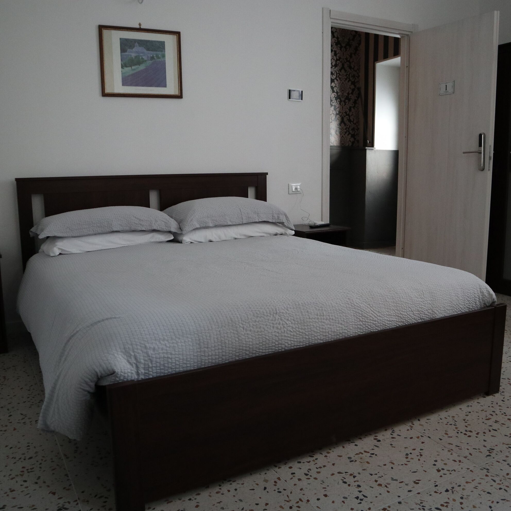 Un dormitorio con una cama grande, una mesita de noche, una puerta y un cuadro enmarcado en la pared.