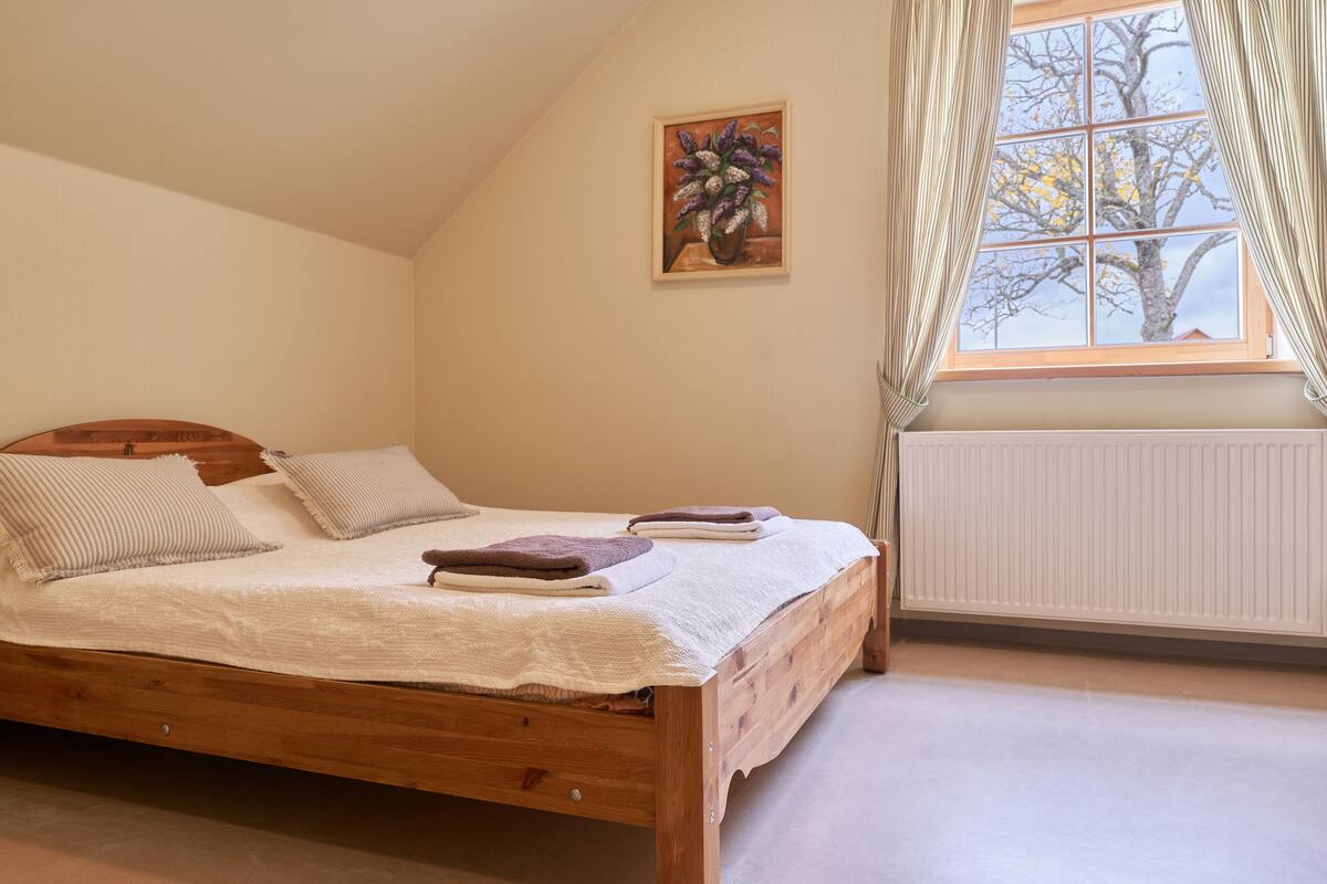 A bedroom with a wooden bed, a window with curtains, and a painting on the wall.