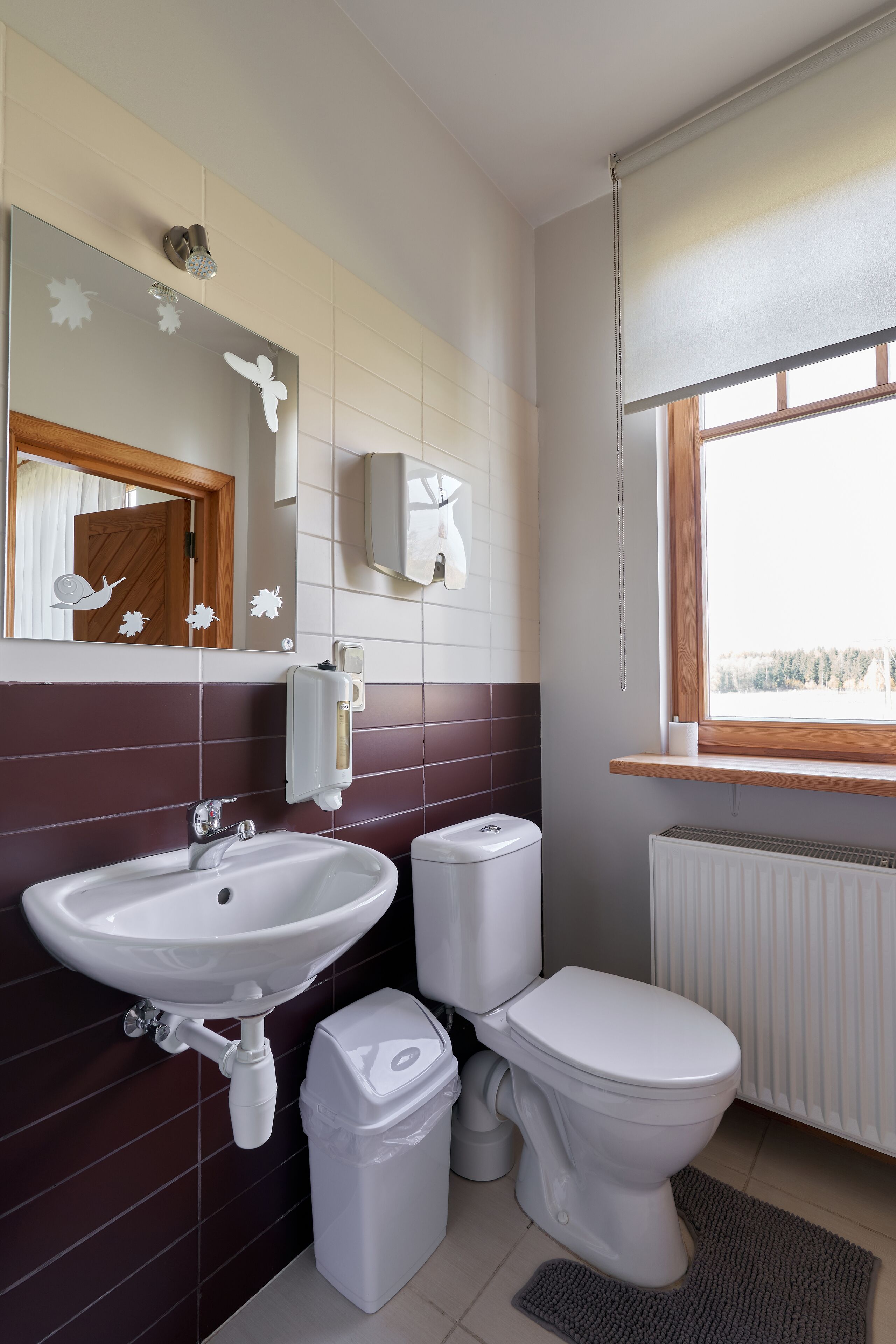 A bathroom with a white sink, toilet, and trash bin, a mirror with decorative stickers, a window with a view, and a wall-mounted soap dispenser.