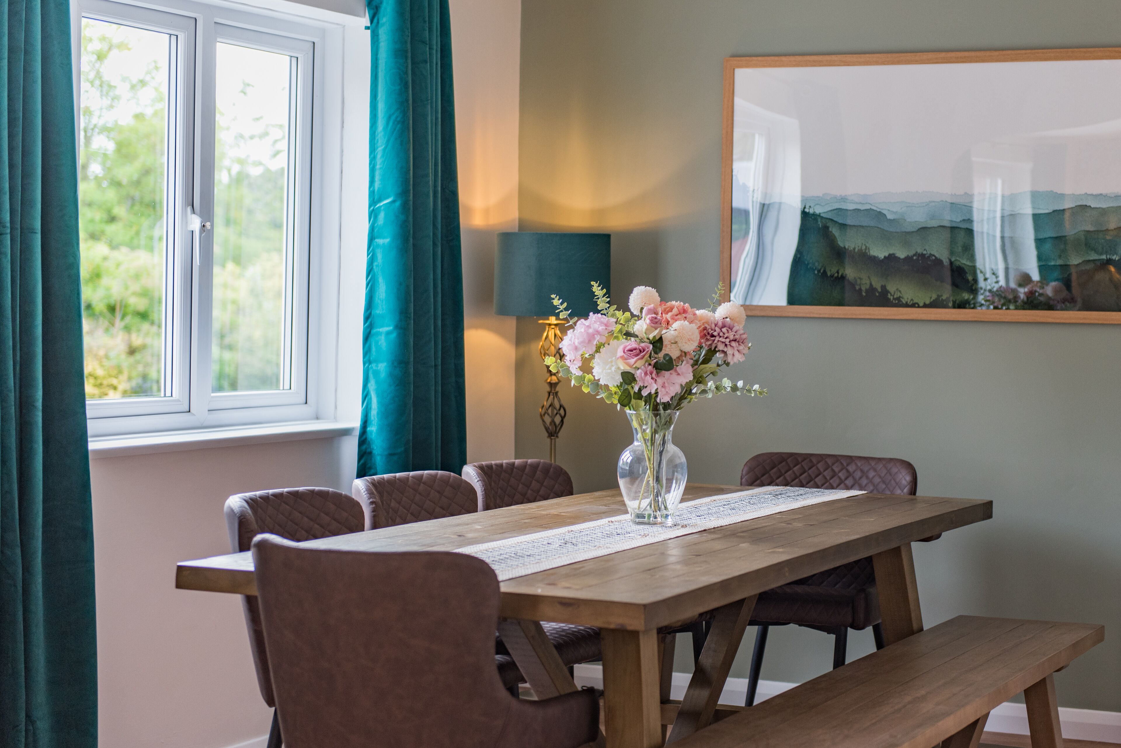 Een eethoek met een houten tafel, comfortabele stoelen, een vaas met bloemen, een lamp en een schilderij aan de muur.