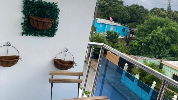 Un balcón con mesa de madera y barandilla metálica, con vistas a una piscina de azulejos azules y un paisaje verde exuberante.