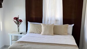 Un dormitorio con cama, una mesita de noche, un jarrón con flores, un ventilador de techo y un aire acondicionado.